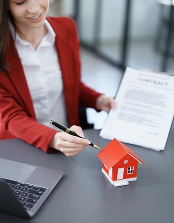 A woman in a red jacket points to a red model house with a pen while holding a contract..