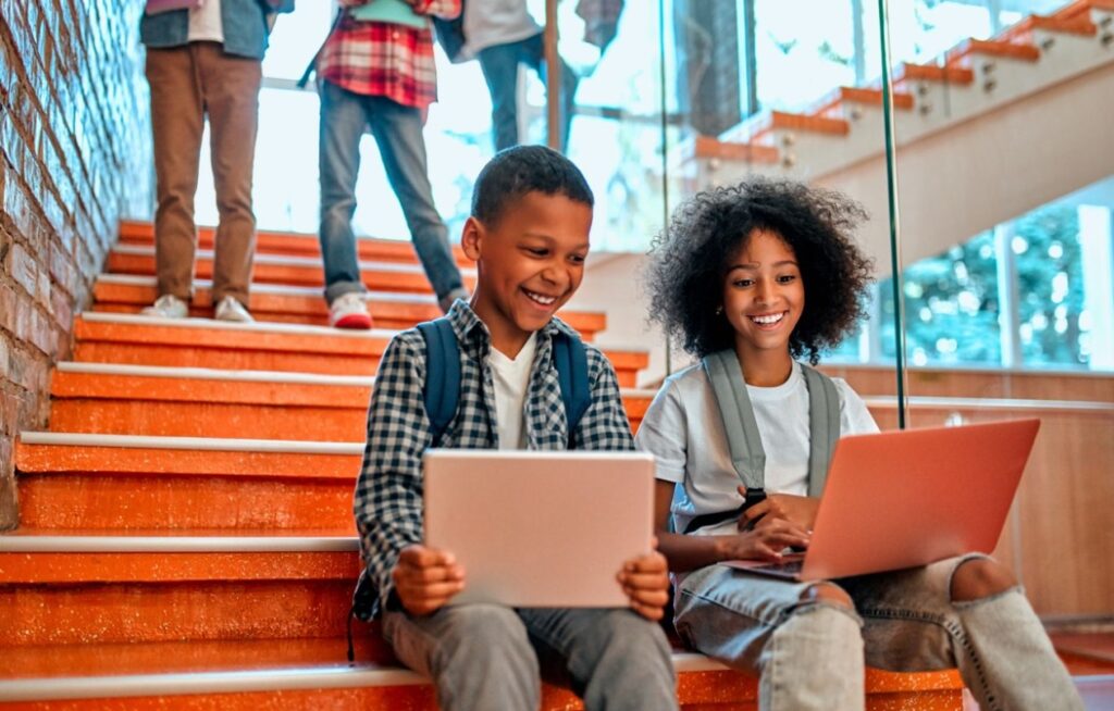 two students looks at laptops while sitting on the steps in their school