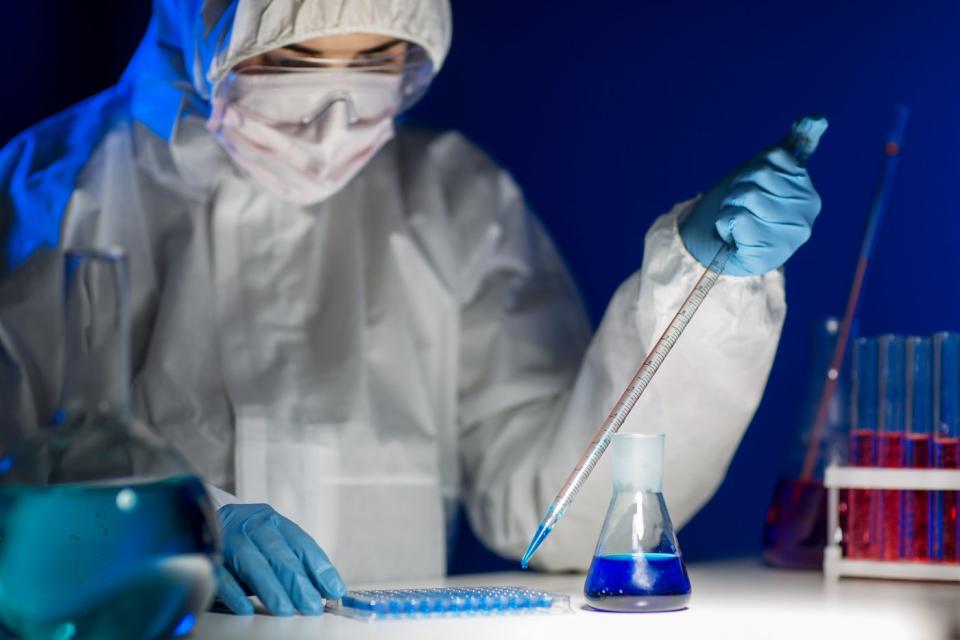 A lab technician wearing gloves and a full-body coverall who's using a pipette to place liquid into a test tray. 