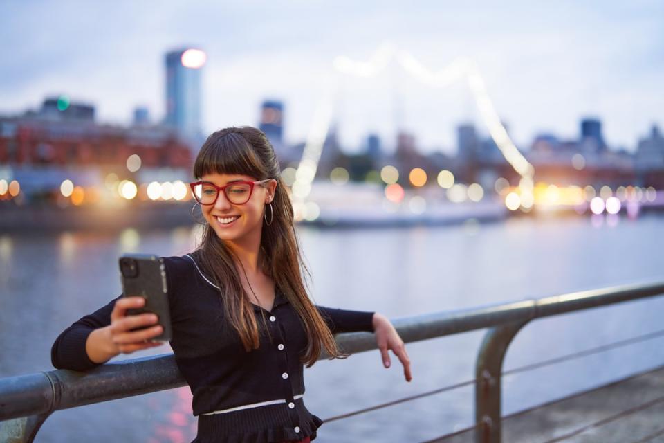 A person smiling while looking at a phone next to a river in an urban setting.