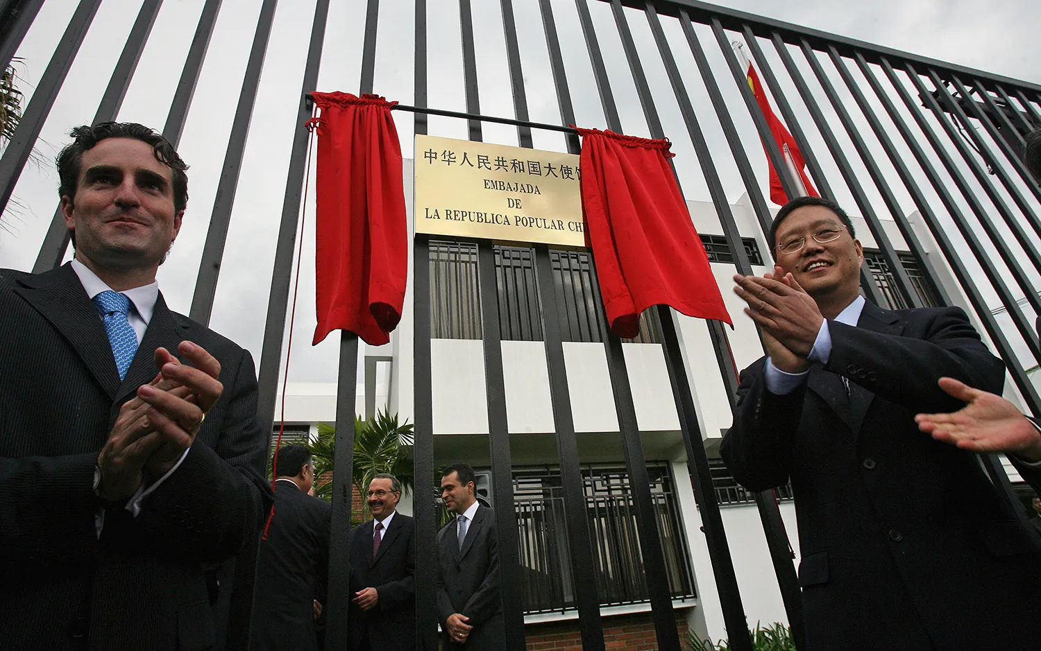 Two men wearing suits clap as they stand in front of a high barred fence. A gold placard with word in Chinese and Spanish hands on the fence with a red curtain parted to reveal it. A building and three other men are seen behind the fence.