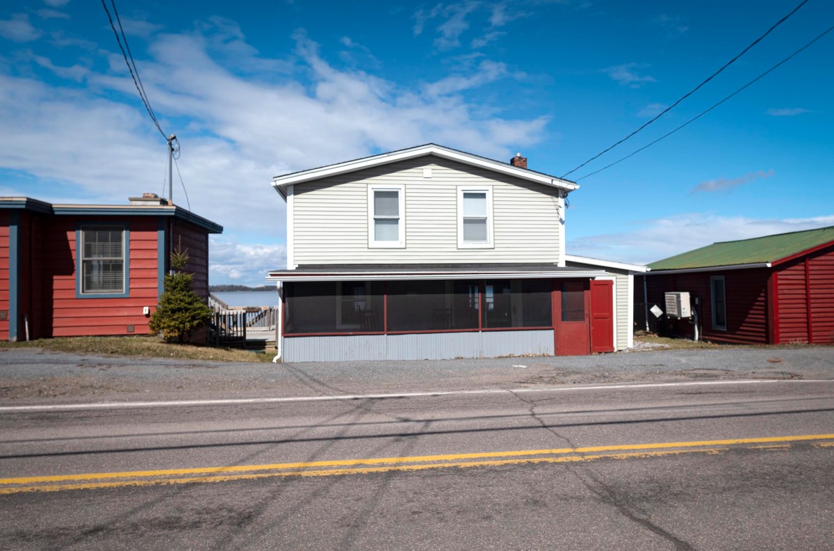 A two-story house with light siding and a red door, flanked by two other houses, is situated along a road under a partly cloudy sky.