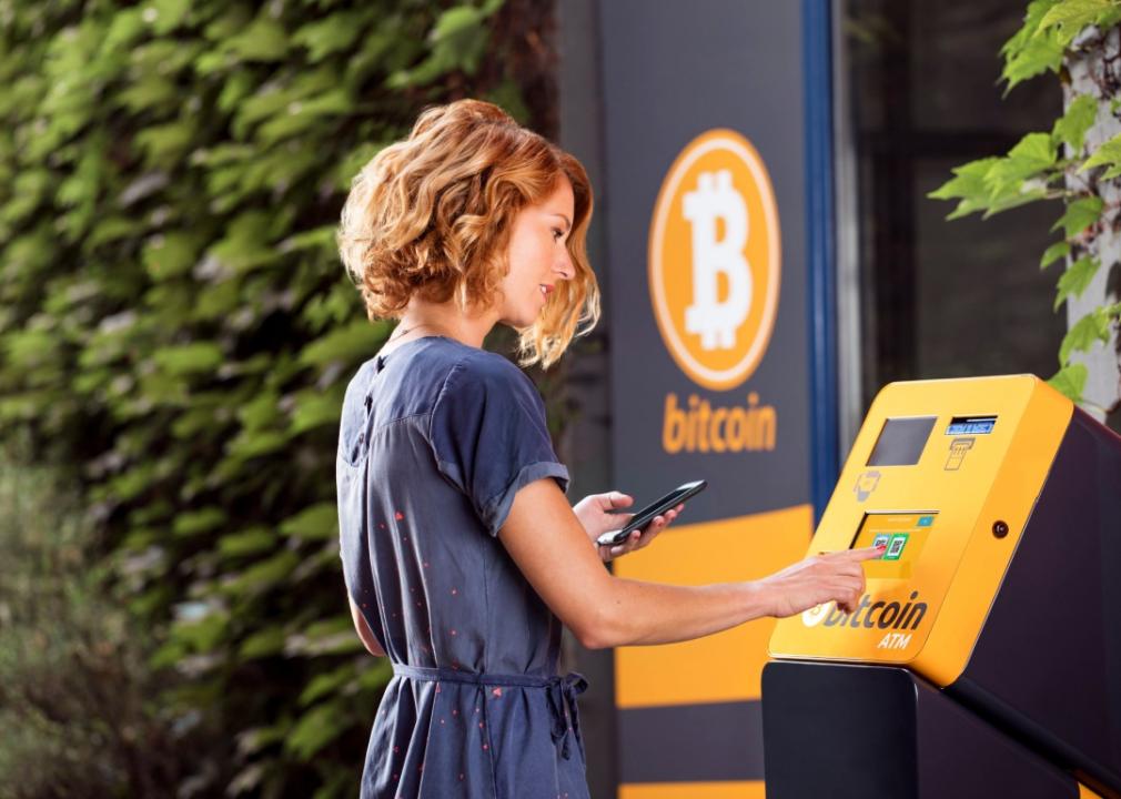 A woman standing at a bitcoin ATM, looking at the screen and using her right hand to press a button, while holding a smartphone in her left hand. 