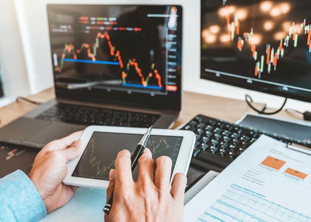 A close up of person's hands analyzing financial data on a tablet, with multiple monitors displaying stock market charts in the background.