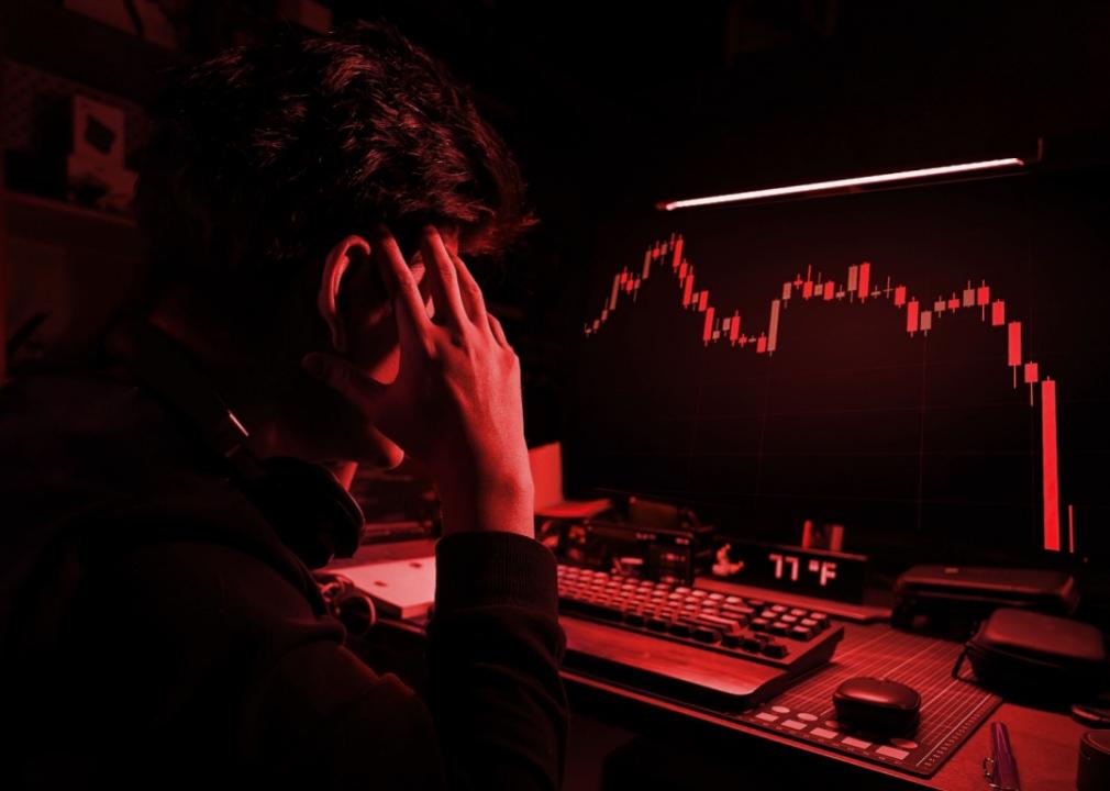 A person sits in a red dimly lit room, hands on their head, staring at a computer screen displaying a downward-trending red graph.