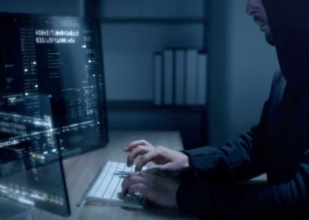 A person in a dark hoodie working in front of two computer monitors in a dimly lit room, their hands illuminated by the screens as they type on the keyboard.