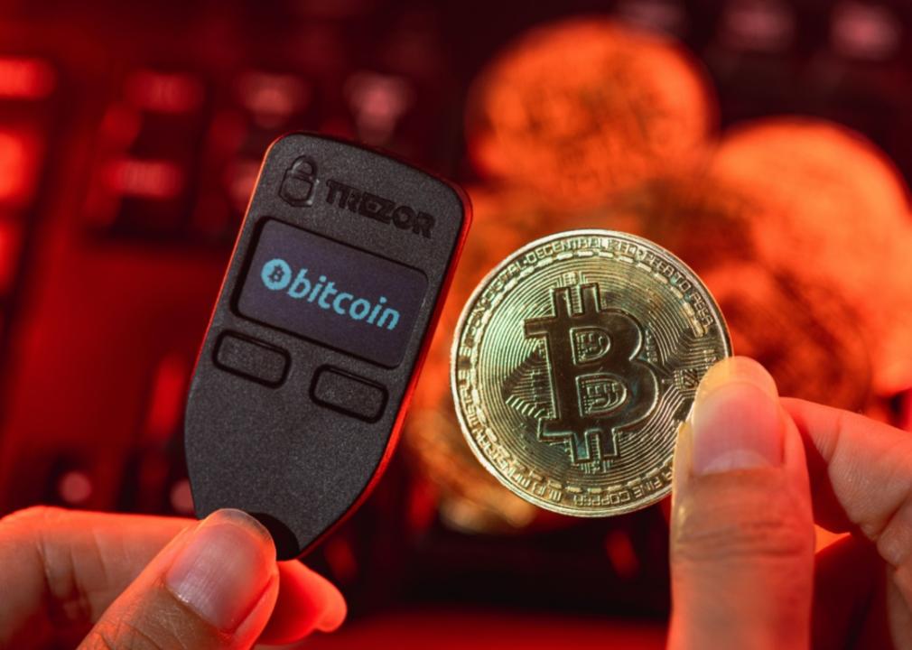 A person's hand holding a Trezor hardware wallet and a gold Bitcoin.