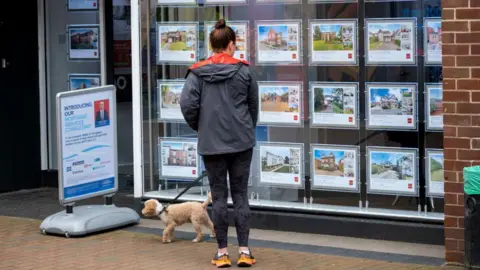 Getty Images Woman with a dog stands in front of an estate agents window looking at adverts for homes.