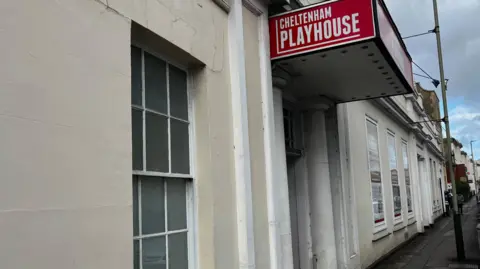 BBC The side of a theatre building. The walls are painted a cream colour. There is a red sign above the door saying Cheltenham Playhouse.