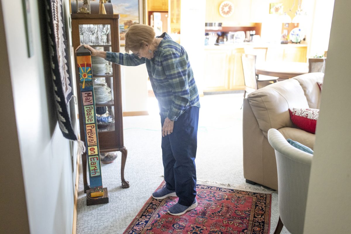 A woman wearing a blue plaid shirt and blue slacks looks at a "peace post" in her home that reads "may peace prevail on earth." She lost money in a cryptocurrency scam