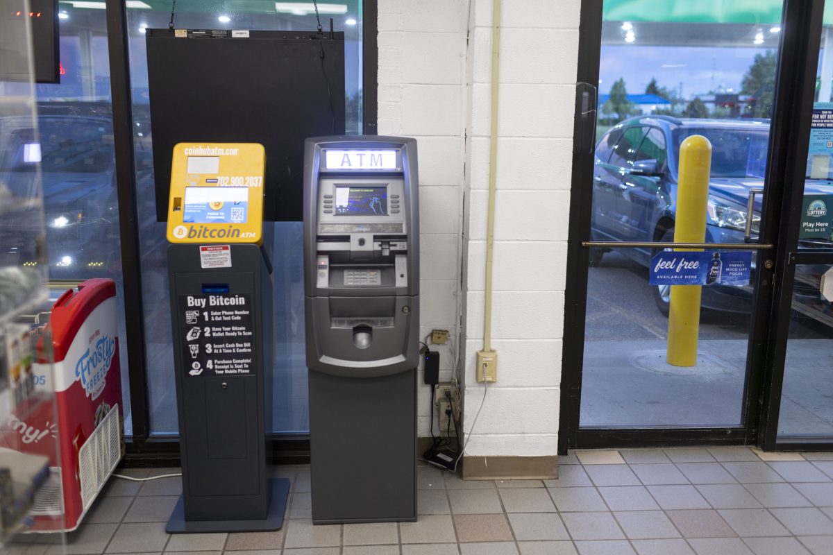 Two ATMs stand next to each other just inside the door of a convenience store. The one on the left is yellow and advertises the ability to buy bitcoin. The one on the right is gray and is a conventional cash machine.