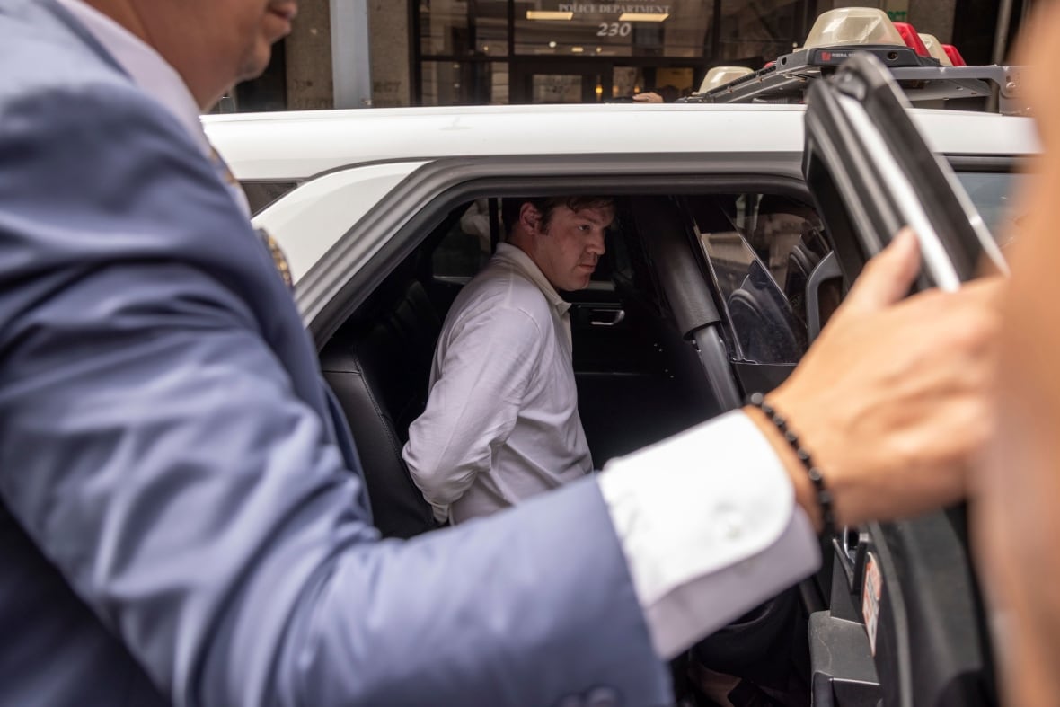A cleanshaven Caucasian man is shown in closeup in a police vehicle.