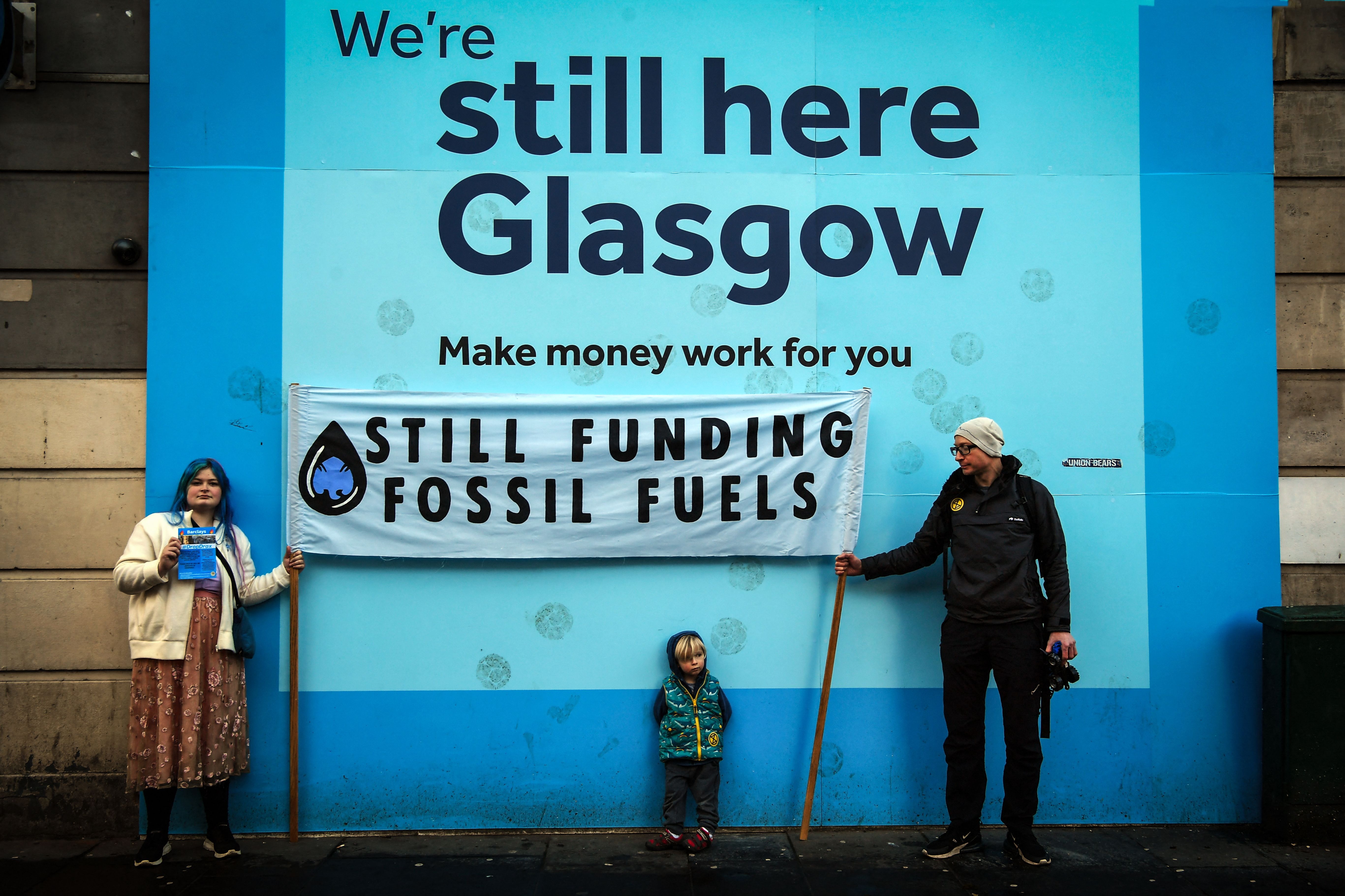 Activists in Glasgow, UK stand in front of a Barclays advert holding a banner that reads “still funding fossil fuels”