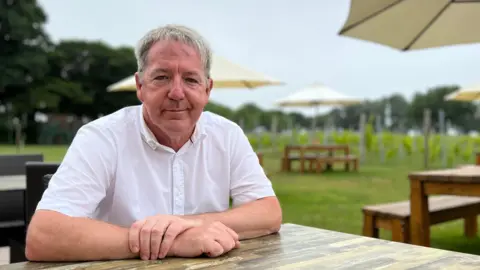 BBC Tim sits at an outdoor wooden table, wearing a white shirt with both arms resting on the surface. The background features large umbrellas shading other tables and benches, set in a grassy area with trees and what appears to be a vineyard in the distance.