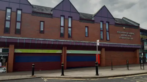 LDRS View of a two-storey retail unit in red brick, with high narrow windows on the first floor. The three units on the ground floor have roller shutters over then, and a sign on the building reads: Houghton-le-Spring Library and Learning Centre.