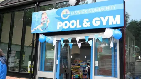 Newcastle City Council Elswick Community Pool and Gym. The glass fronted building has a large blue sign hanging above its door. Balloons and bunting are displayed above the entrance to celebrate its reopening.