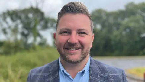 Sam Kurtz smiles at the camera. He has brown eyes and short dark hair and a beard, and is wearing a grey plaid blazer and light blue shirt. Trees and grass can be seen blurred behind him.