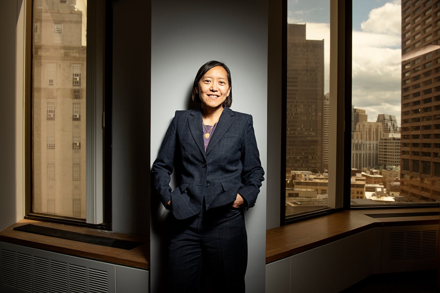A portrait of Yvonne Hao standing against a wall in an office setting with windows on either side of her showing some office buildings in downtown Boston behind her.