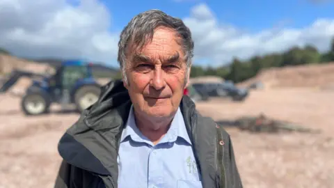 A man in a blue shirt and green jacket looks at the camera in front of a blue tractor and the remnants of a small fire.