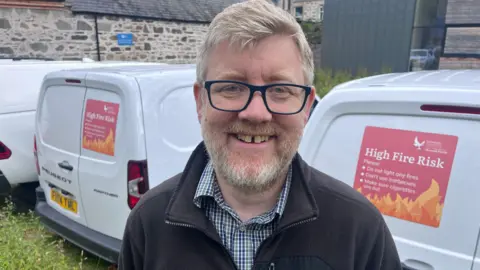 A mark with dark glasses and a beard, wearing a fleece and checked shirt, stands in front of two white vans displaying warnings of a high fire risk.