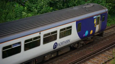 PA Media Landscape shot of a Northern-branded train. There are hedgerows behind and the train is moving away from the photographer.